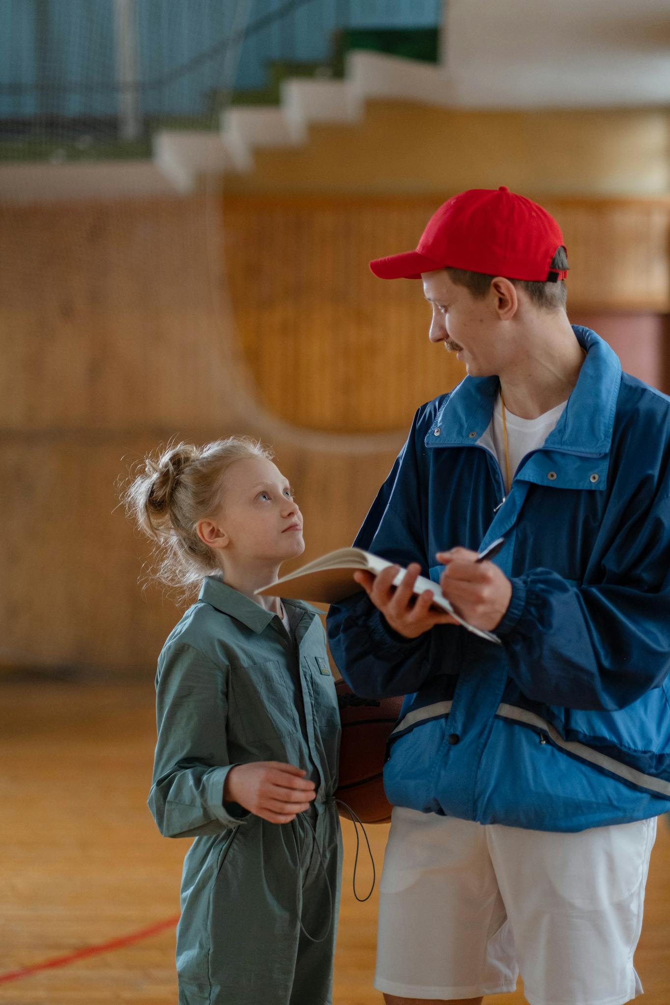 A young coach with a clipboard guides a child athlete during a training session indoors.